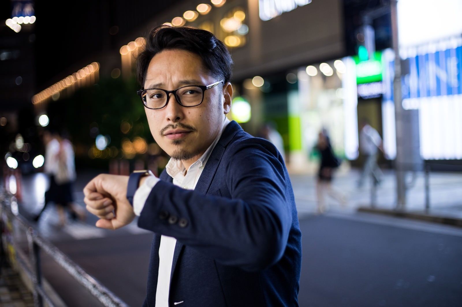 夜間の繁華街で腕時計を確認する眼鏡のビジネスマン［モデル：ゆうせい］