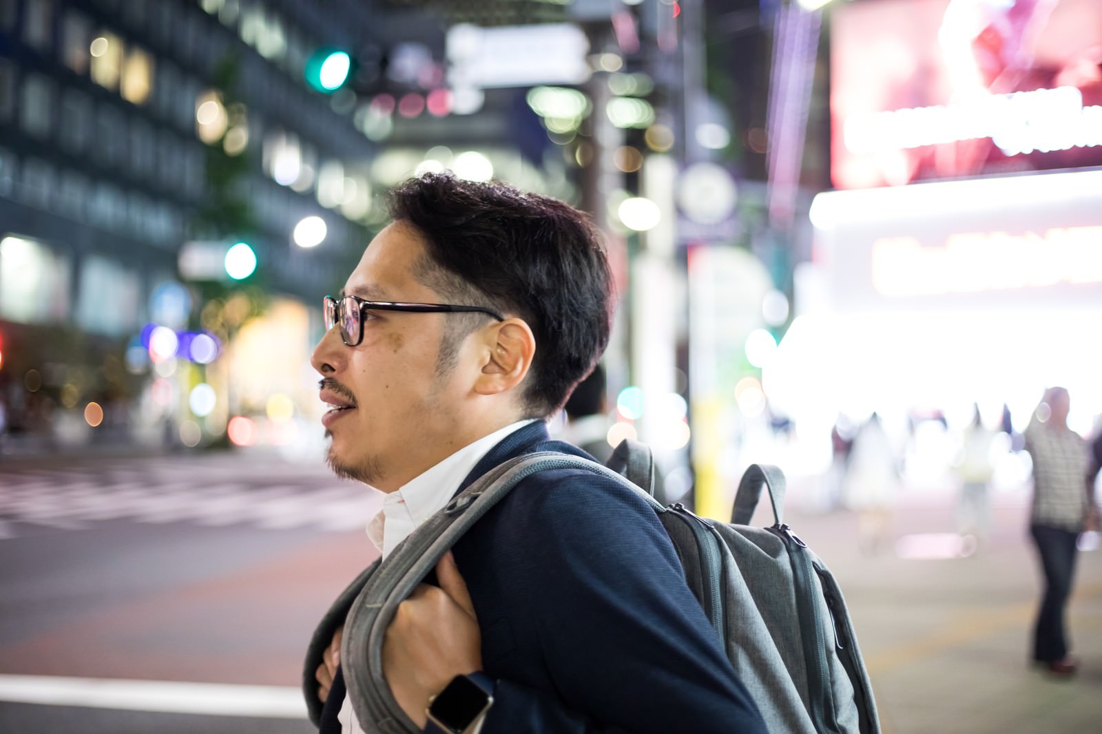 夜の繁華街でネオンを背景に歩く眼鏡をかけたスーツ姿の会社員男性［モデル：ゆうせい］