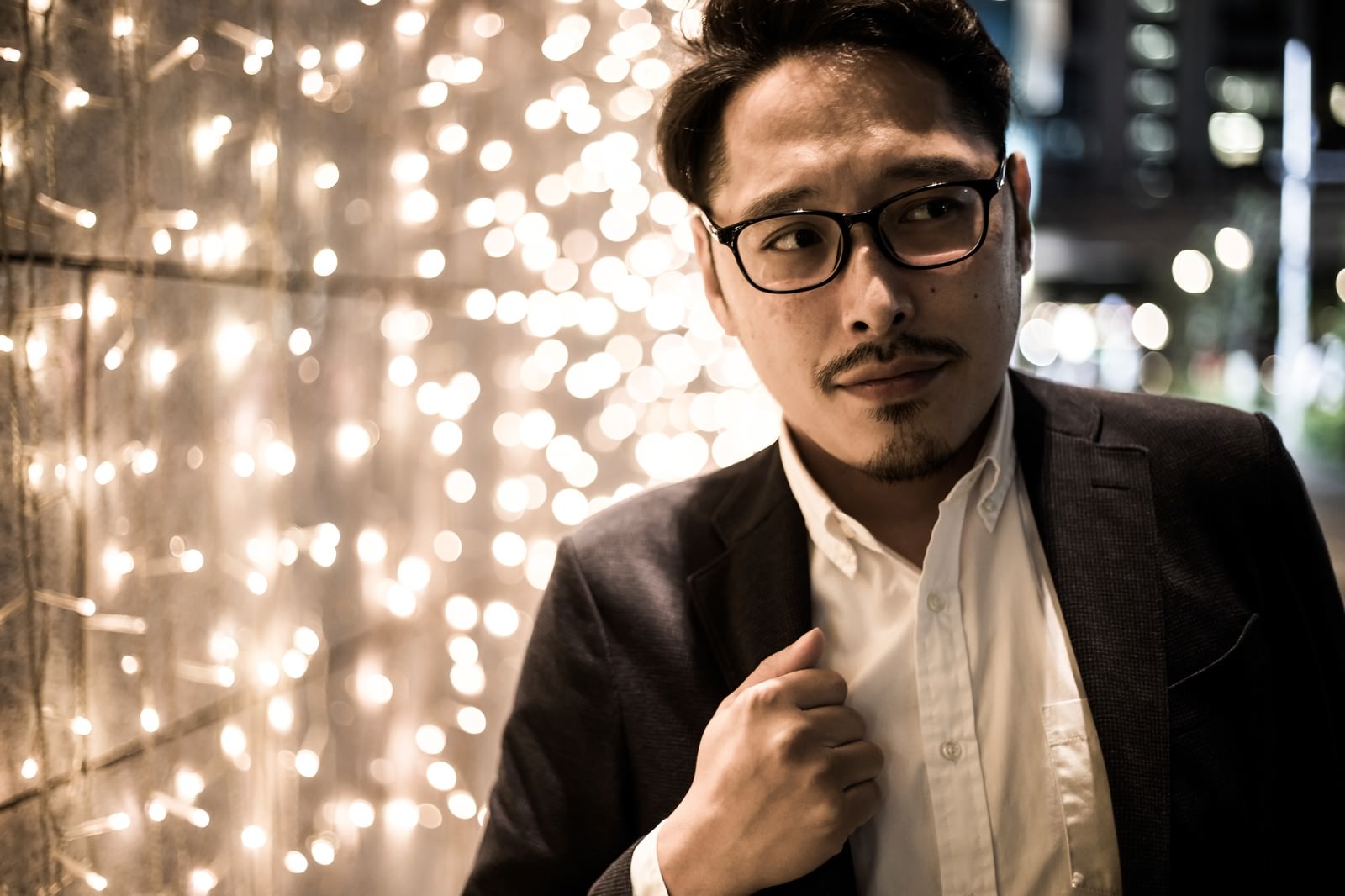 イルミネーションが輝く夜の背景でネクタイを整えるスーツ姿の眼鏡男性［モデル：ゆうせい］