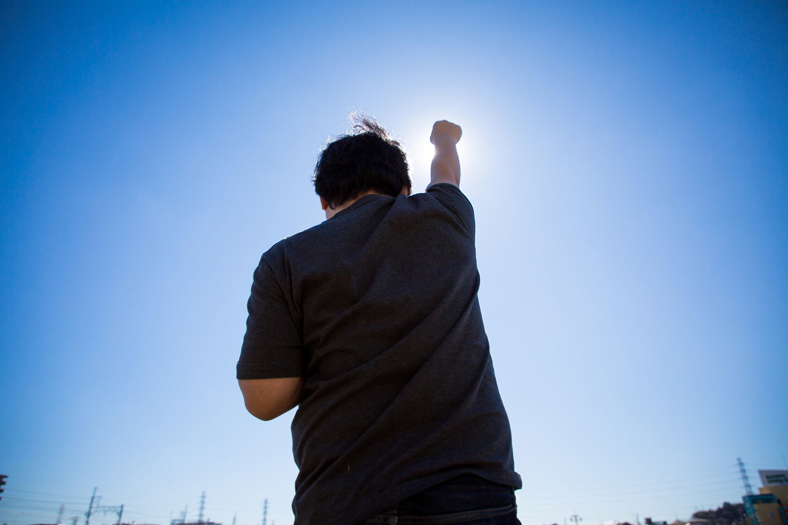 太陽に向かって拳を高々とあげる男性の後ろ姿、逆光で青空をバックに［モデル：段田隼人］