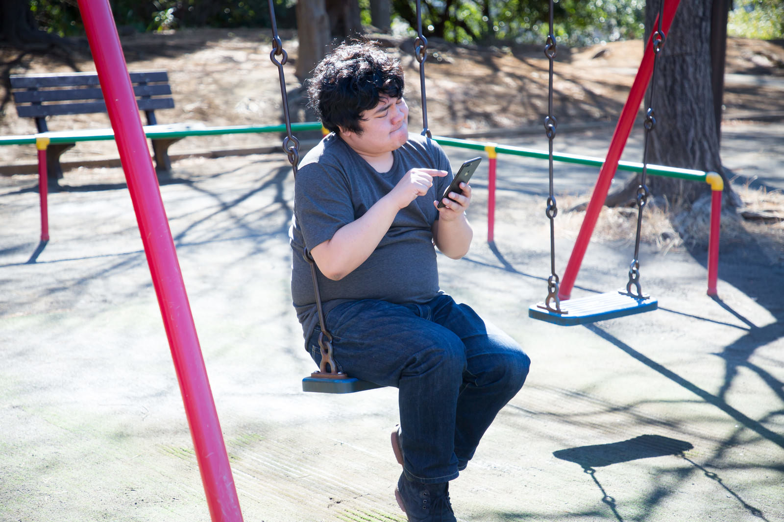 公園のブランコに座ってスマートフォンを操作する男性［モデル：段田隼人］