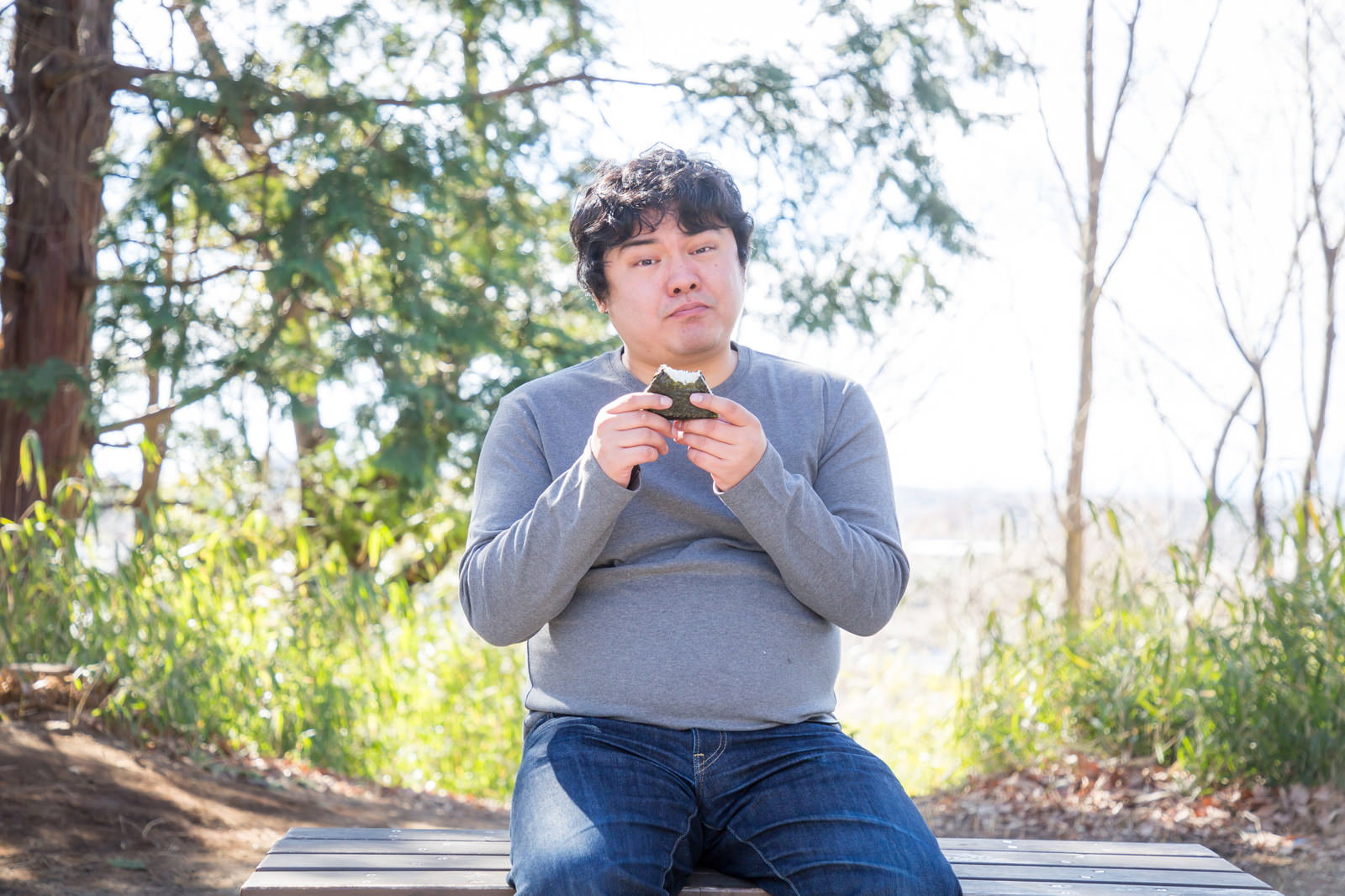 屋外のベンチでおにぎりを両手で持ち食べている男性。背景は樹木と曇り空。［モデル：段田隼人］