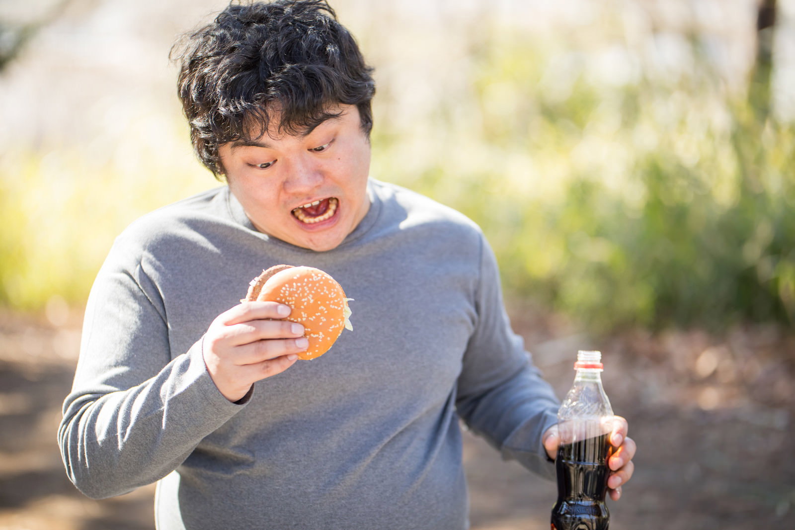 グレーのパーカーを着た男性が屋外でハンバーガーを食べながらコーラボトルを持つ［モデル：段田隼人］