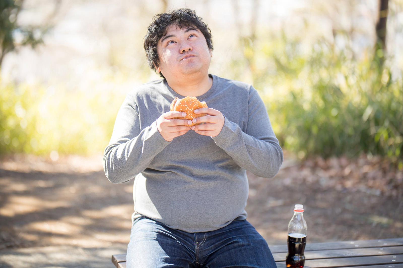 屋外のベンチでハンバーガーを両手で持って食べる男性［モデル：段田隼人］