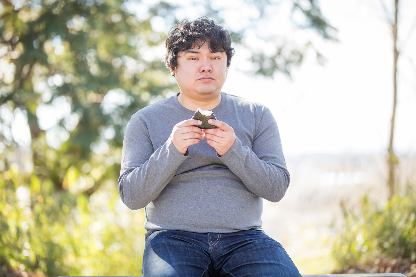 屋外のベンチでおにぎりを食べる肥満体型の男性［モデル：段田隼人］