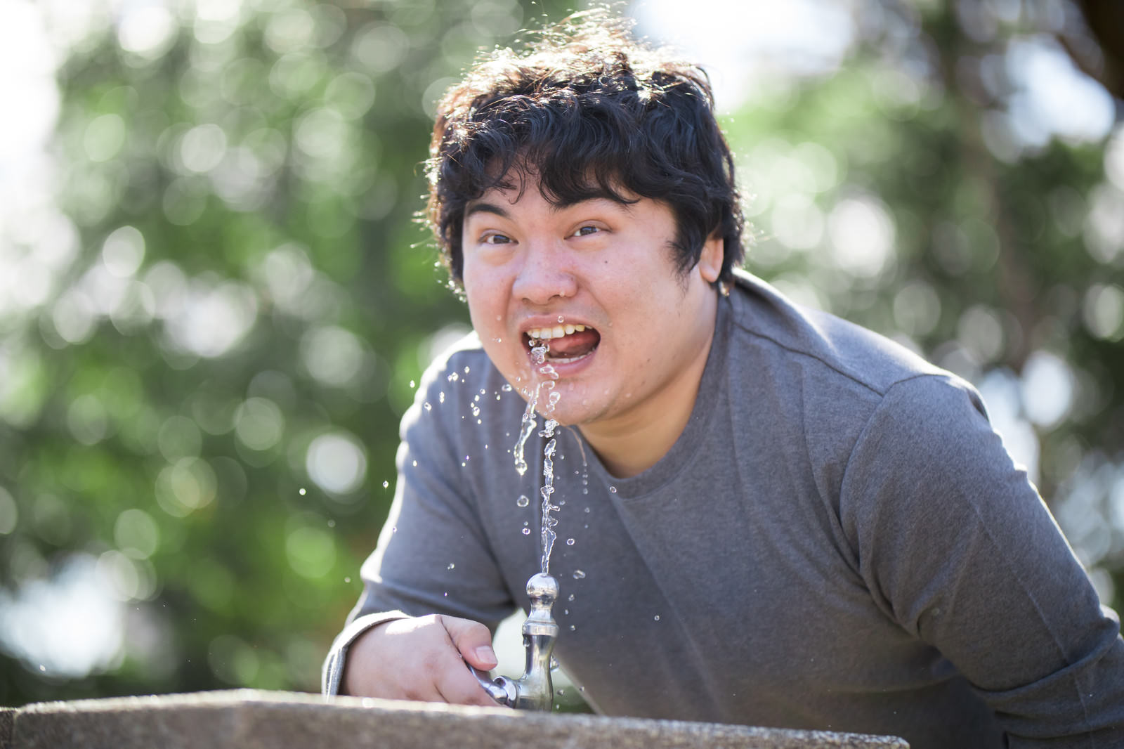 公園の水飲み場で水を飲むグレーのTシャツを着た男性［モデル：段田隼人］