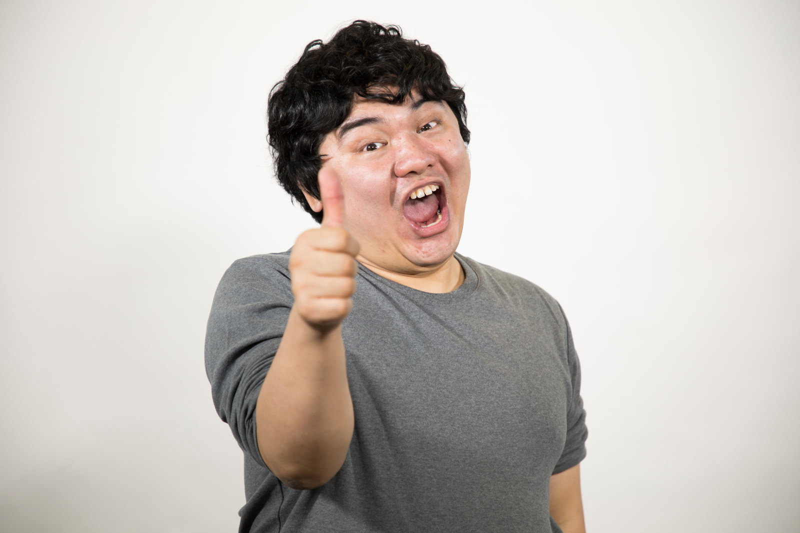 白い壁の前でサムズアップして笑う男性［モデル：段田隼人］