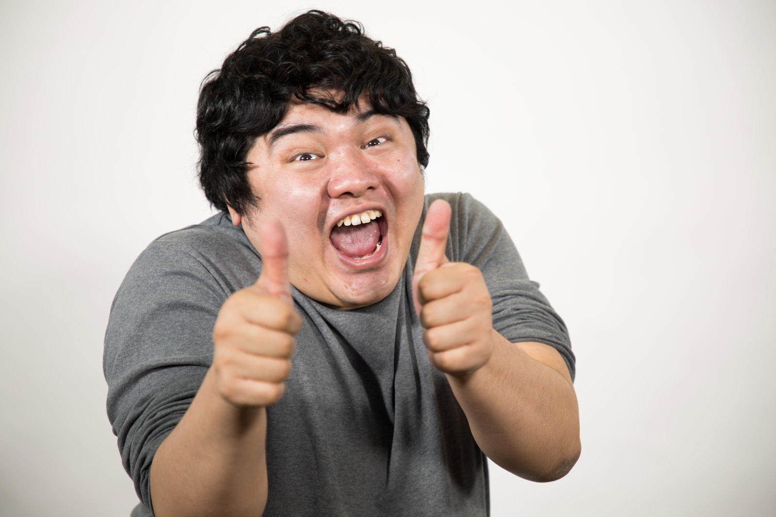 白い壁の前で両手サムズアップして笑う男性［モデル：段田隼人］