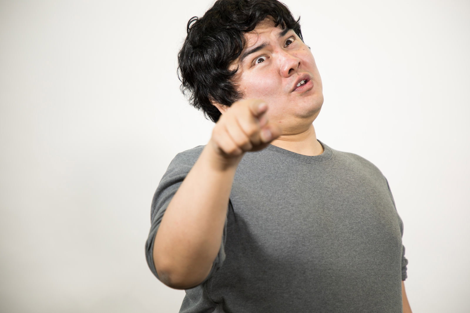 グレーのTシャツの男性が上を向き、右手の人差し指で指さしながら怒りの表情を見せている［モデル：段田隼人］