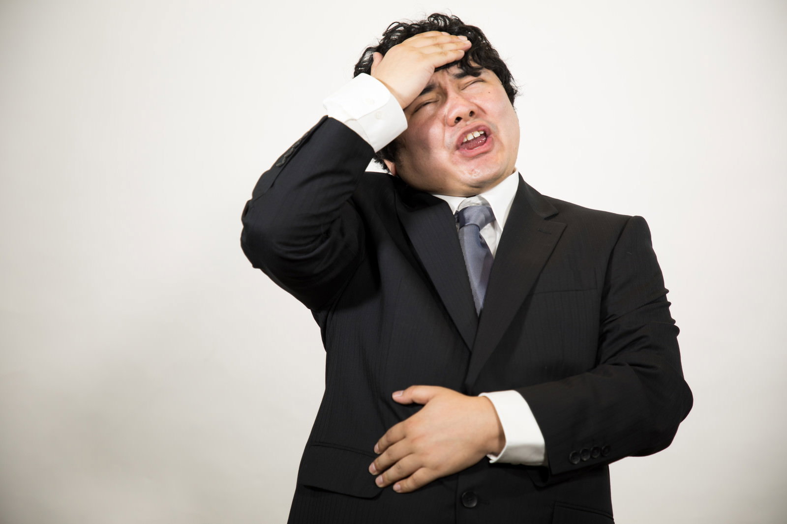 頭を抱えて苦痛な表情を浮かべる黒いスーツ姿の男性［モデル：段田隼人］