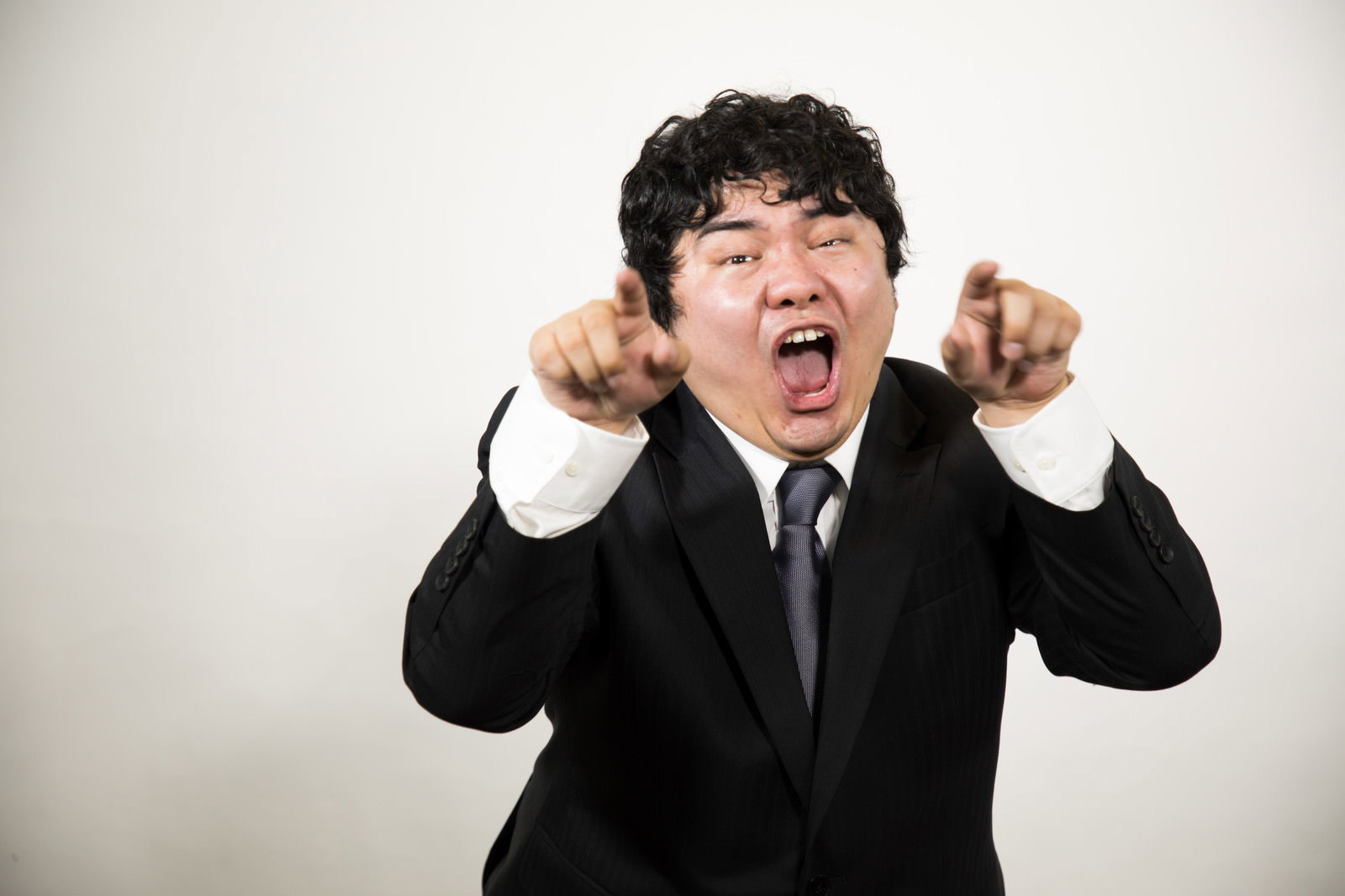 黒いスーツを着た男性が両手の人差し指を突き出して指をさし、笑顔で挑発的なジェスチャーをしている［モデル：段田隼人］