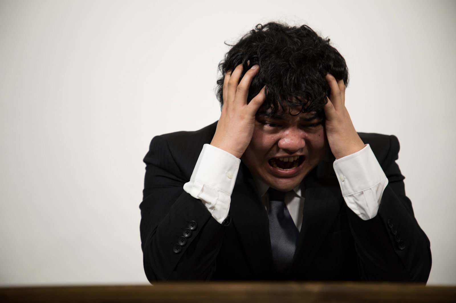 机に向かい両手で頭を抱える黒いスーツ姿の男性［モデル：段田隼人］
