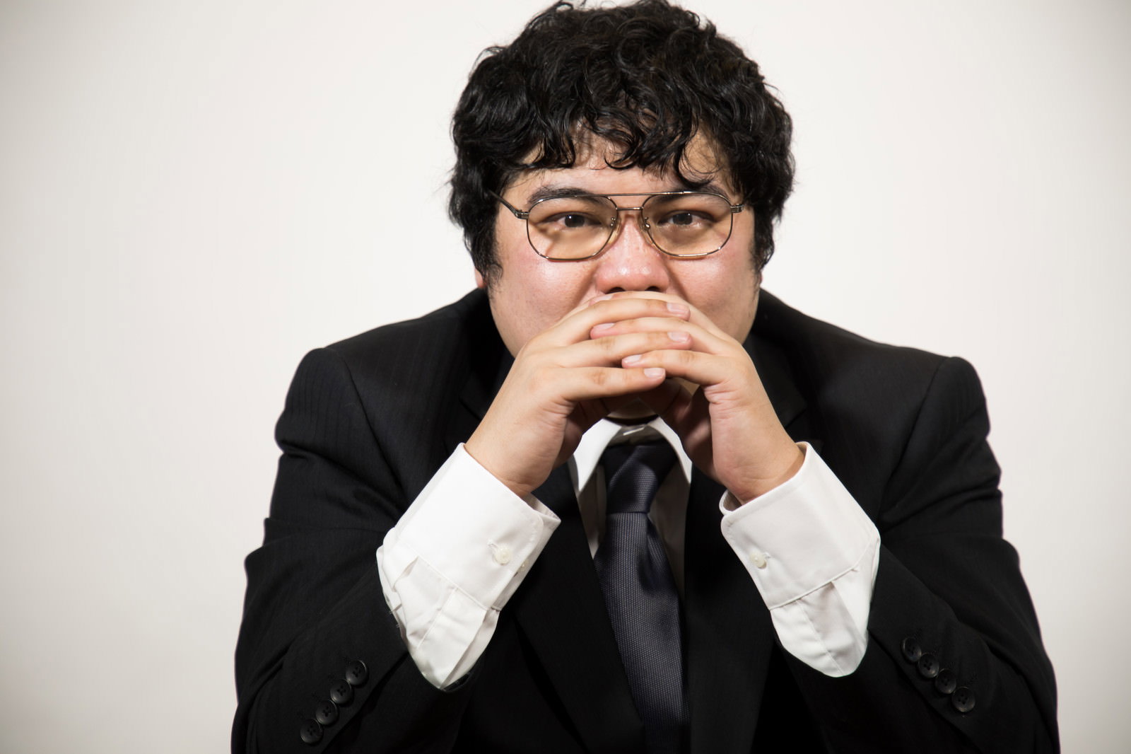 黒いスーツを着て眼鏡をかけた男性が両手を組んで困った表情で考え込んでいる［モデル：段田隼人］