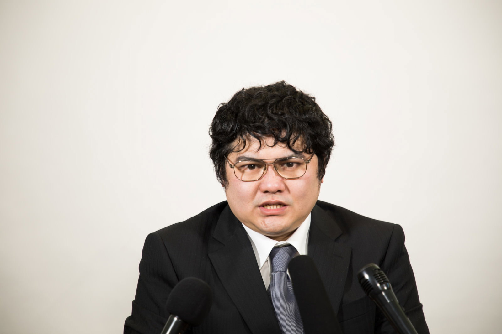黒いスーツと眼鏡の男性役員がマイクの前で険しい顔で記者会見に対応している［モデル：段田隼人］