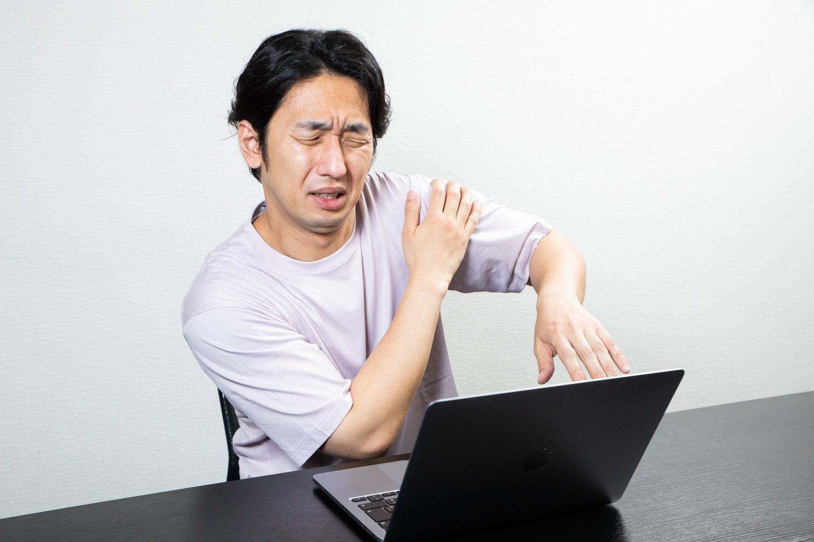 白いシャツを着た男性がデスクでノートパソコンを使いながら腕の違和感を訴えている様子［モデル：大川竜弥］