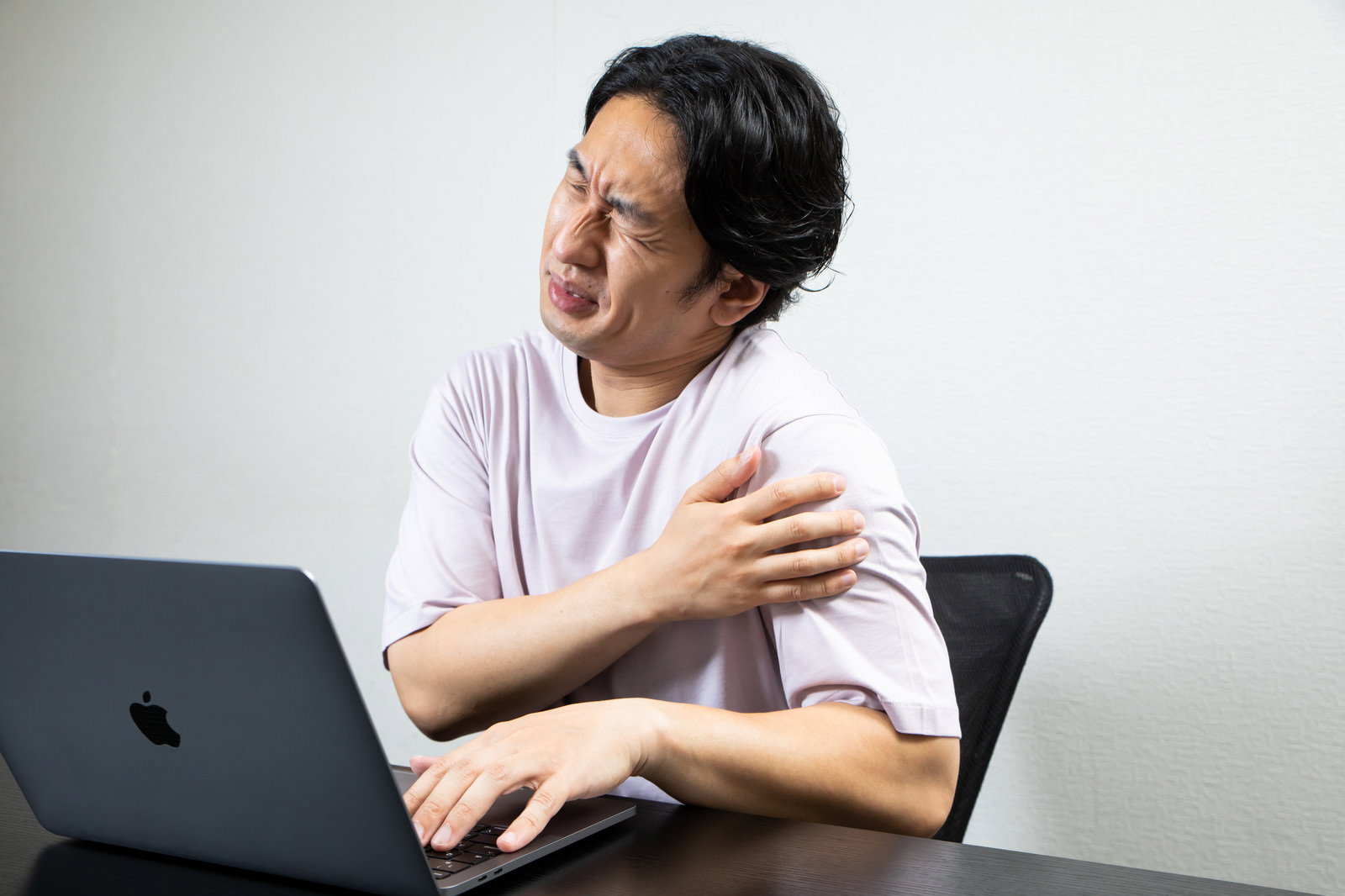ノートパソコンの前で肩に手を当て、しかめっ面をする白シャツの男性［モデル：大川竜弥］