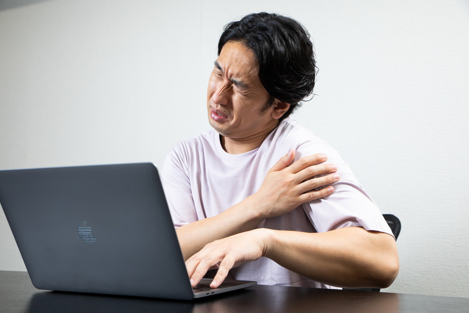 ノートパソコンの前で肩に手を当て痛みを感じている男性［モデル：大川竜弥］