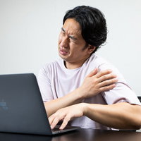 デスクワーク中に肩の痛みを感じてしかめっ面の男性の写真