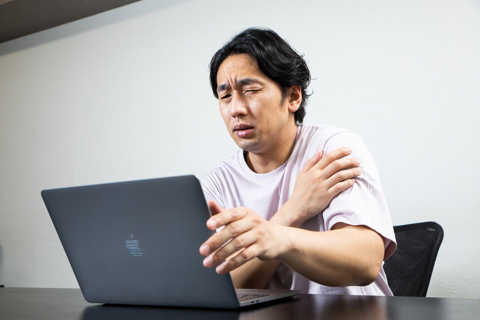 ノートパソコンの前で右肩を押さえて辛そうな表情を見せる白いTシャツの男性［モデル：大川竜弥］