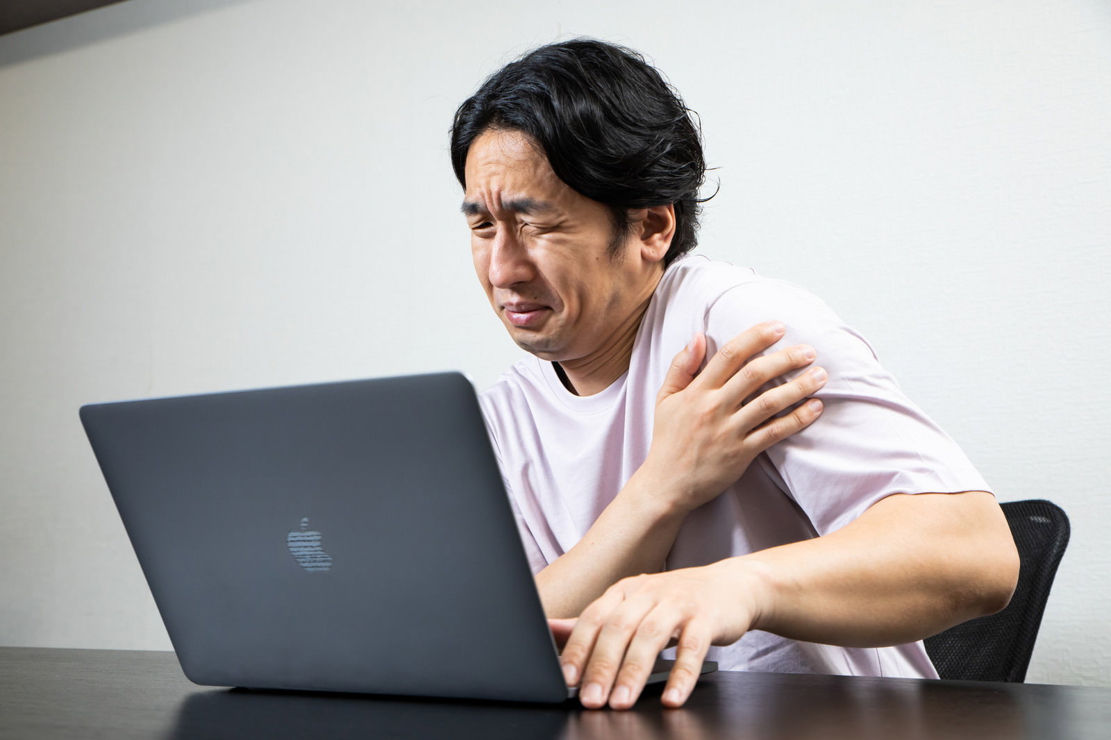 肩の痛みをかばいながらノートパソコンで作業する白いシャツの男性［モデル：大川竜弥］