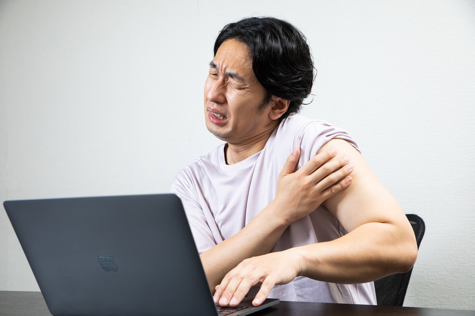 パソコンの前で肩に手を当てて痛そうな表情を浮かべる男性［モデル：大川竜弥］