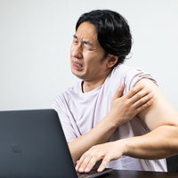 副反応による肩の痛みに苦しむ男性のデスクワーク中の体調不良の写真