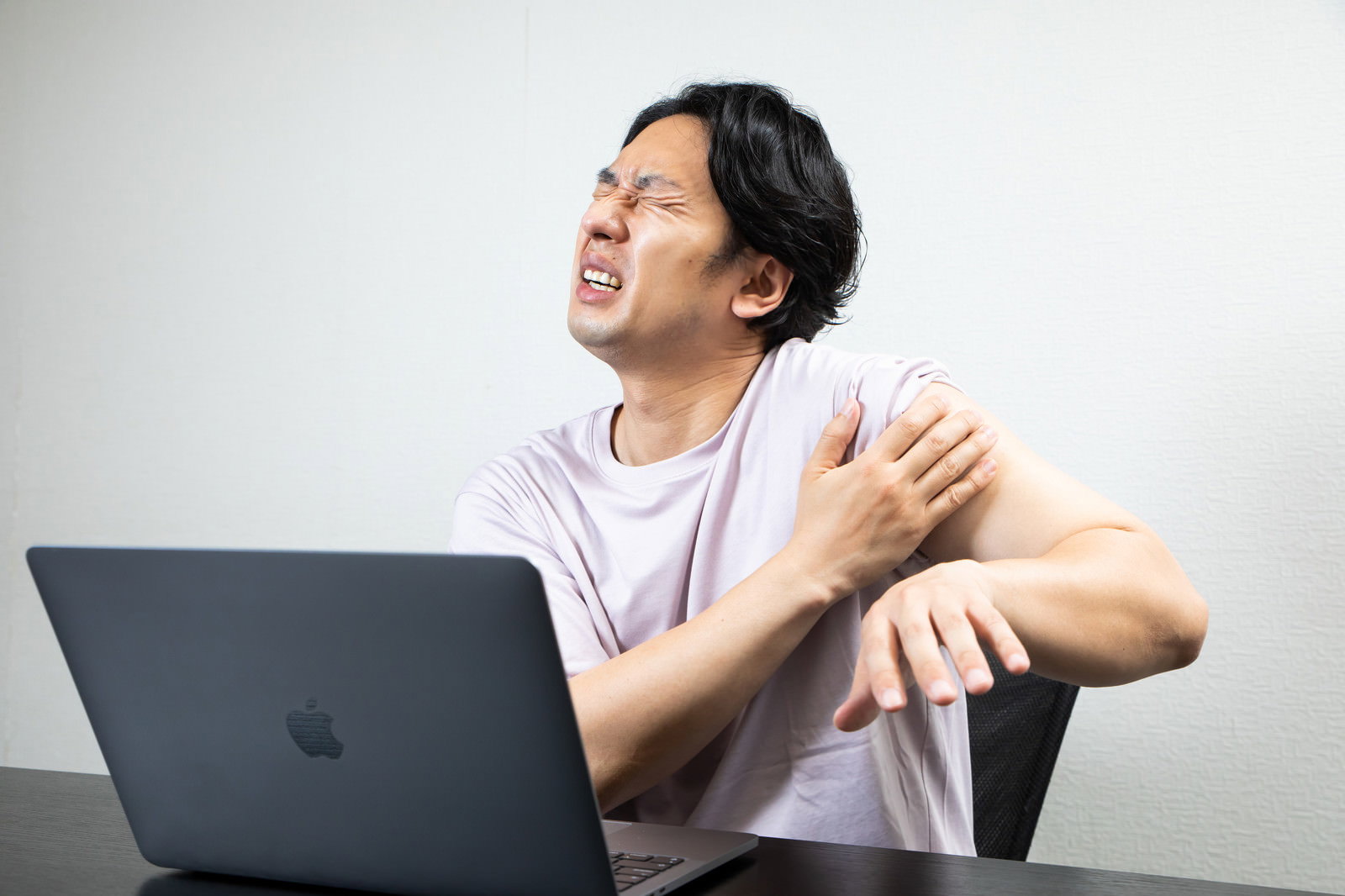 デスクでノートパソコンを前に右肩を押さえて痛がる白いTシャツの男性［モデル：大川竜弥］