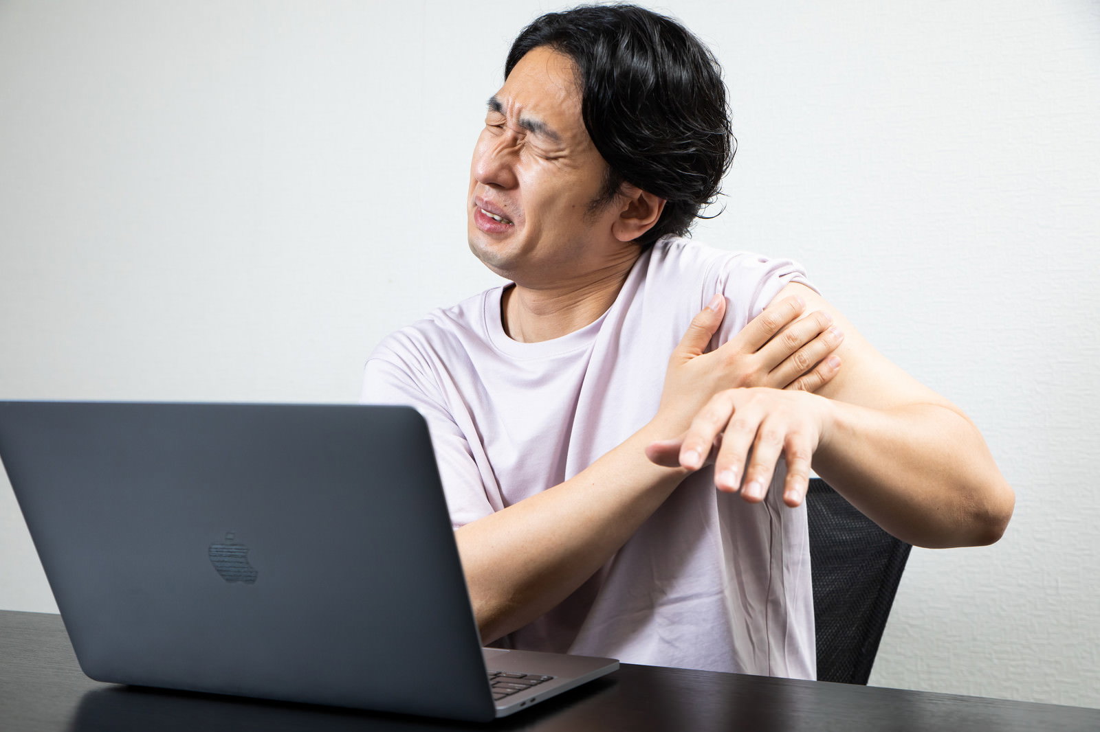 肩こりで痛そうな表情を浮かべながらノートパソコンを使う男性［モデル：大川竜弥］
