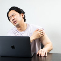 ワクチン接種の副反応で肩が痛む男性がデスクワーク中に肩を抑えるの写真