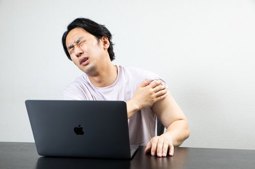 ワクチン接種の副反応で肩が痛む男性がデスクワーク中に肩を抑える