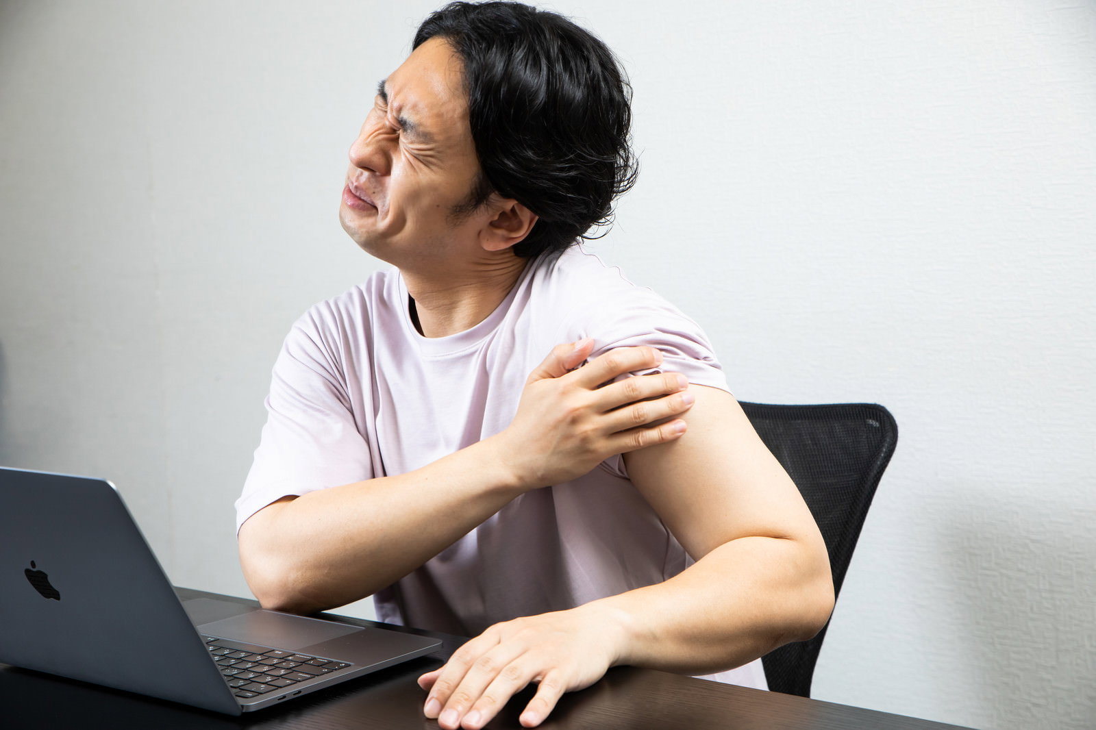 パソコンの前で左肩を押さえて痛がる白いTシャツの男性［モデル：大川竜弥］