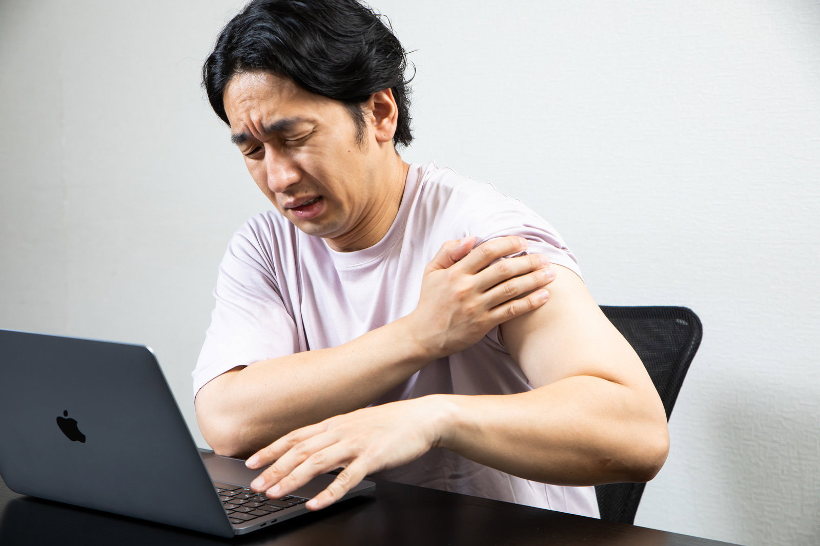 デスクでパソコン作業をしながら肩を押さえて痛がる白いTシャツの男性［モデル：大川竜弥］