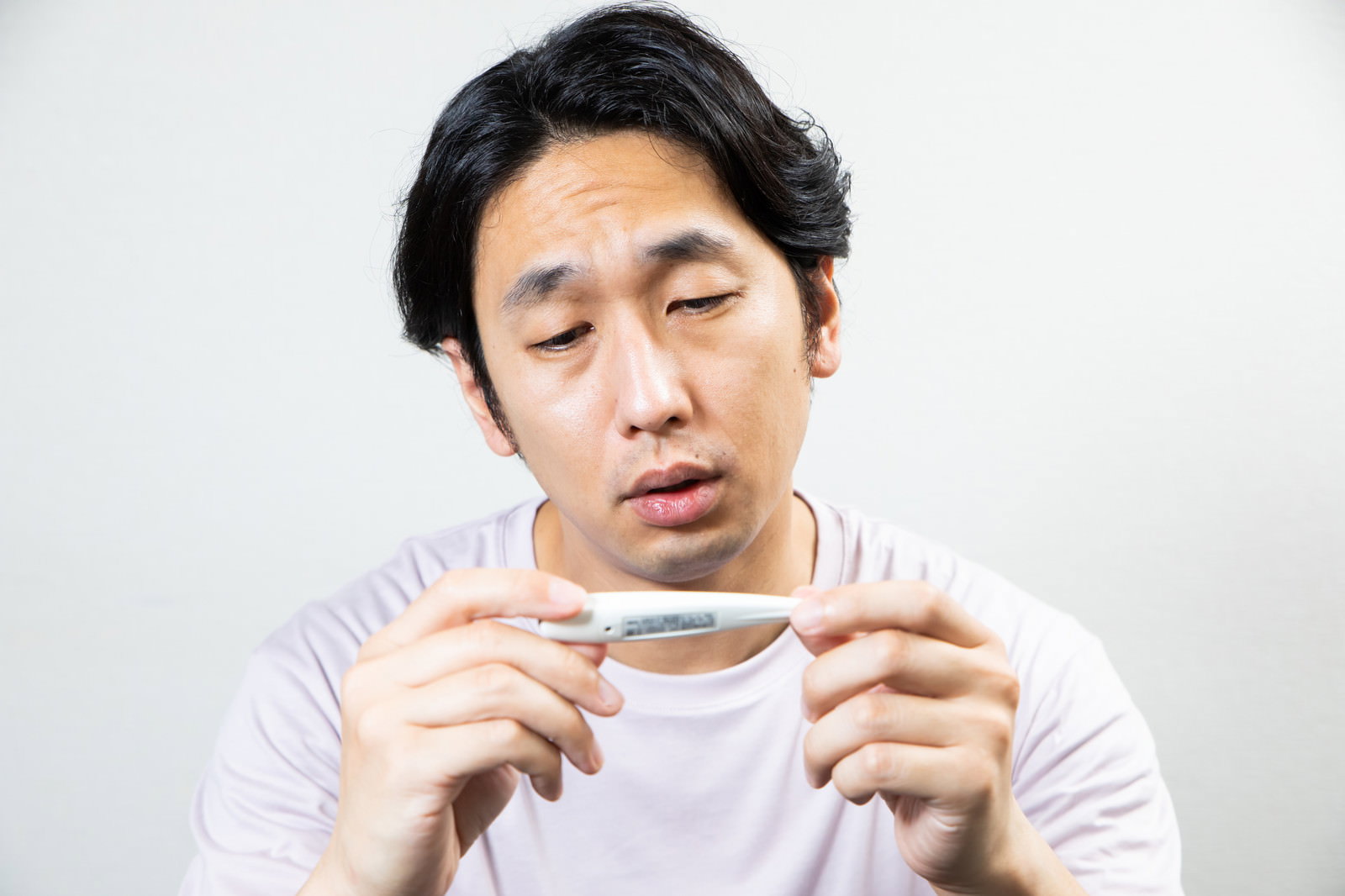 体温計を両手で持ちながらうなだれている白いTシャツの男性［モデル：大川竜弥］