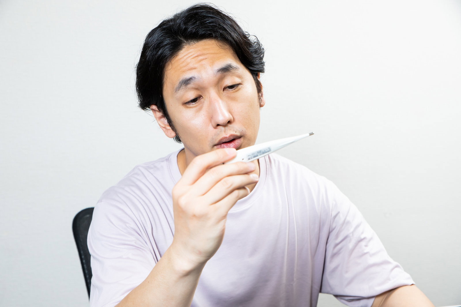 白いTシャツを着た男性が椅子に座り、体温計を手に持ちながらうなだれている［モデル：大川竜弥］