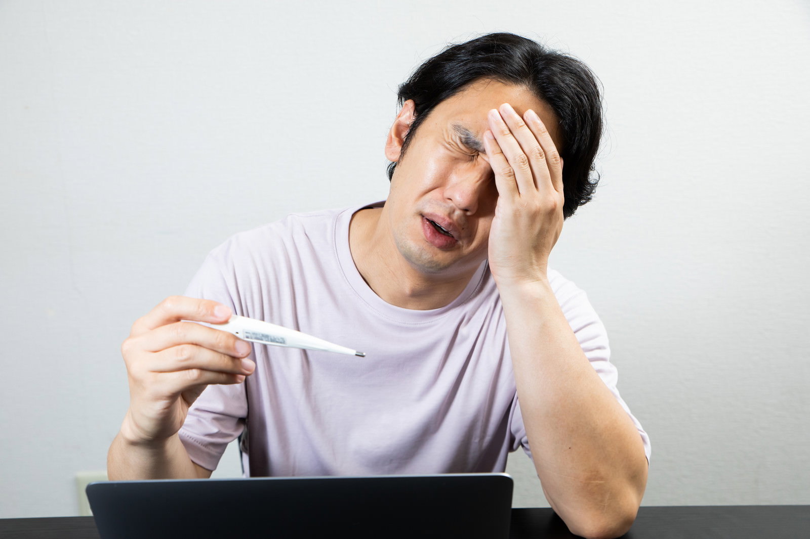 体温計を持ち片手で頭を押さえる白いTシャツの男性［モデル：大川竜弥］