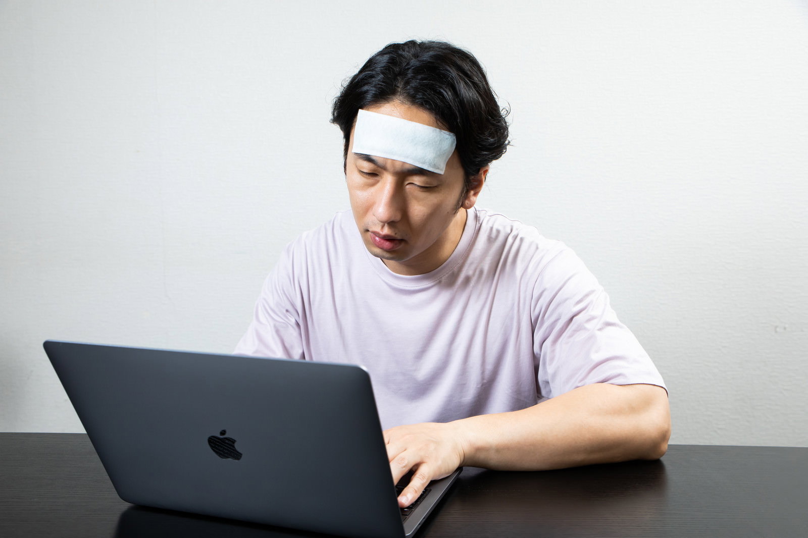 額に冷えピタを貼りノートパソコンの前でぐったりしている男性［モデル：大川竜弥］