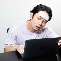 体調不良でパソコンにしがみ付く男性、額に冷えピタを貼った姿の写真