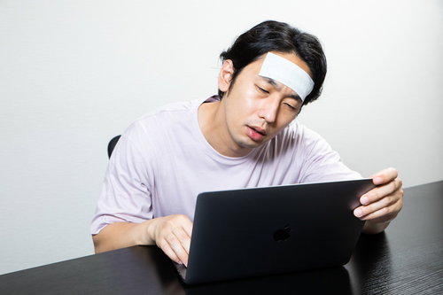 体調不良でパソコンにしがみ付く男性、額に冷えピタを貼った姿