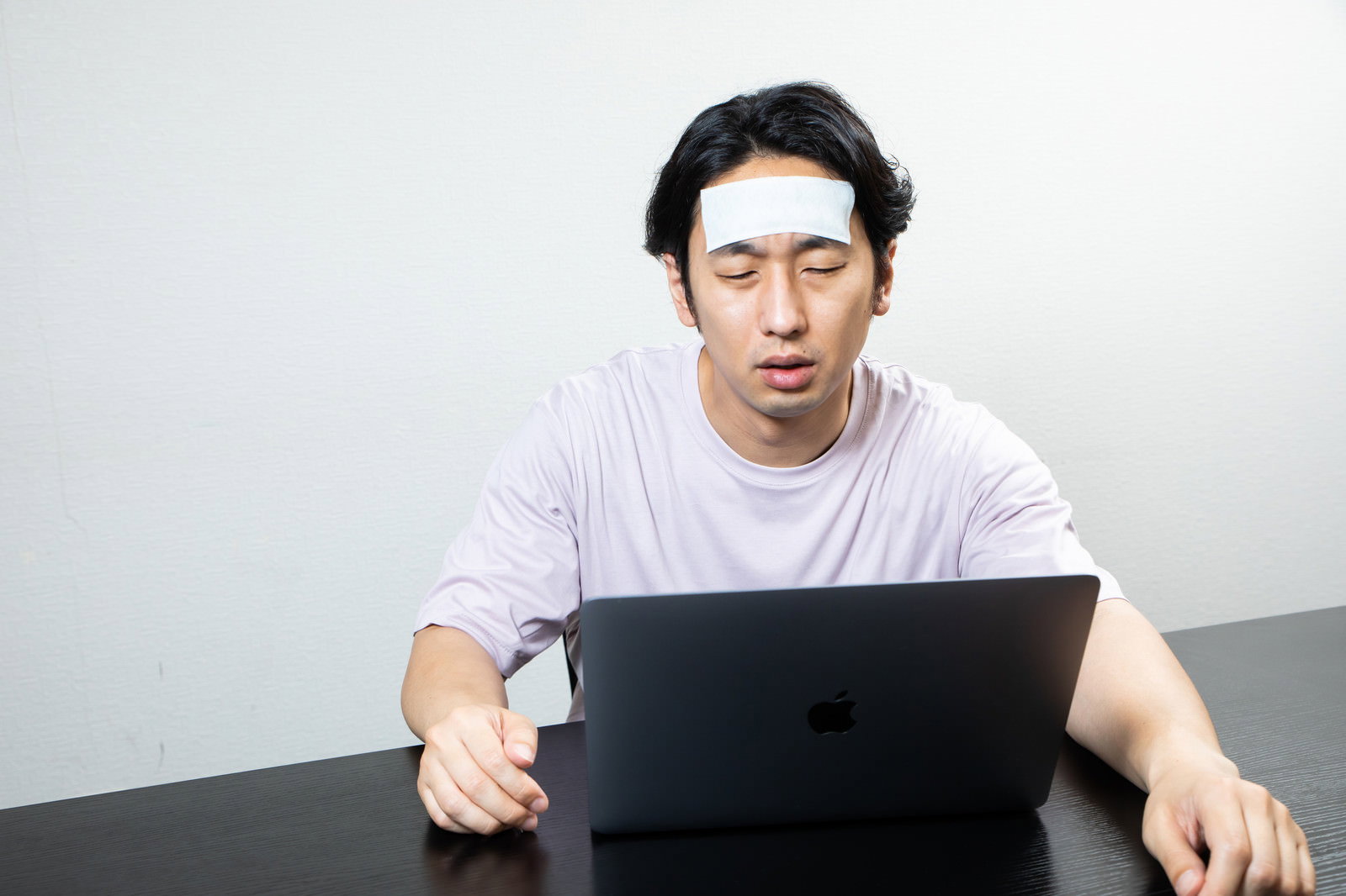 おでこに冷却シートを貼りパソコン作業をする疲れた表情の男性［モデル：大川竜弥］