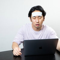 おでこを冷やしてリモートワークする寝不足の男性の写真