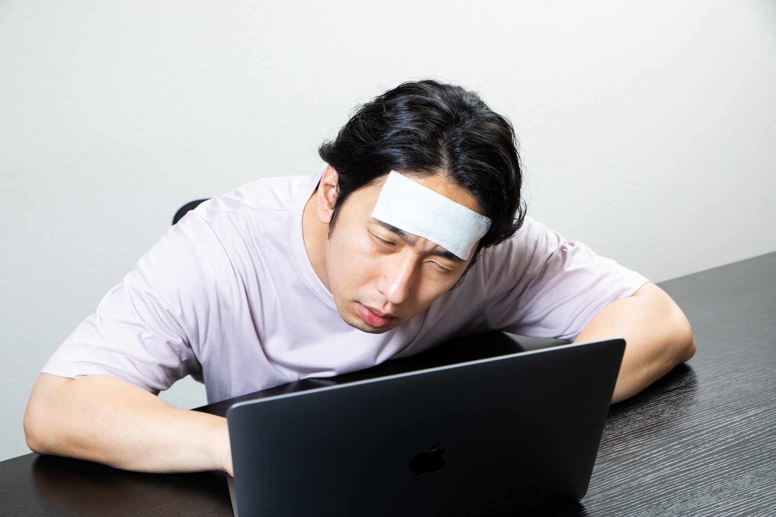おでこに冷却シートを貼り、肘をついてノートパソコンに向かう体調不良の男性［モデル：大川竜弥］