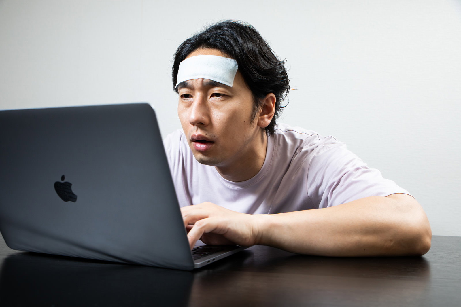 おでこに冷えピタを貼りながらノートパソコンで作業する体調不良の男性［モデル：大川竜弥］