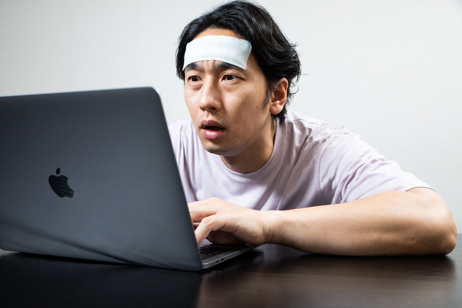 冷えピタを額に貼りながらノートパソコンで作業する男性［モデル：大川竜弥］