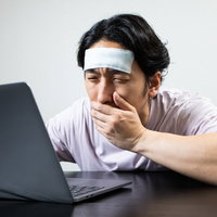 PCの前で吐き気を感じる働き過ぎの男性、おでこに冷えピタを貼り疲労困憊の写真
