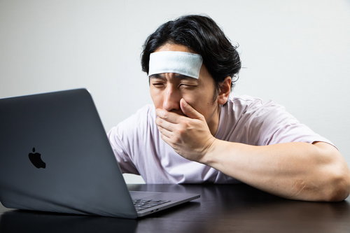 PCの前で吐き気を感じる働き過ぎの男性、おでこに冷えピタを貼り疲労困憊