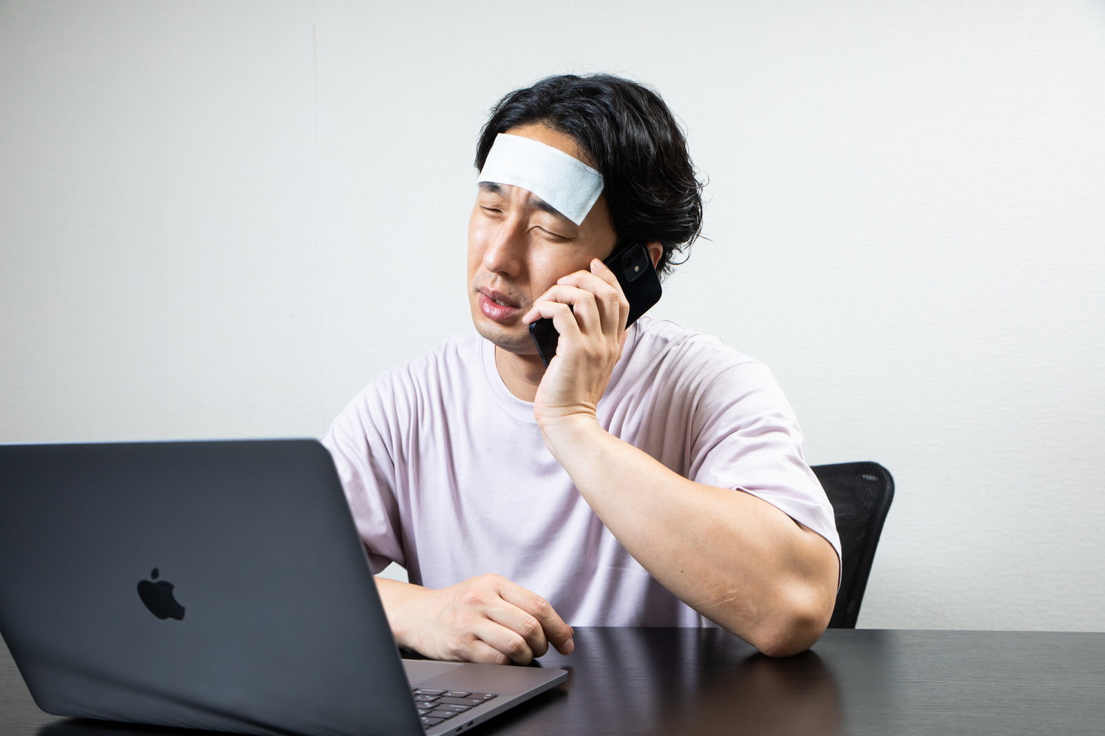 おでこに冷却シートを貼ってパソコンの前で電話をする男性［モデル：大川竜弥］