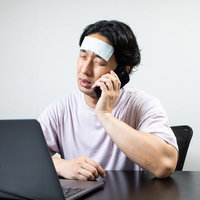 おでこを冷やしながら欠勤連絡する在宅勤務の男性の写真