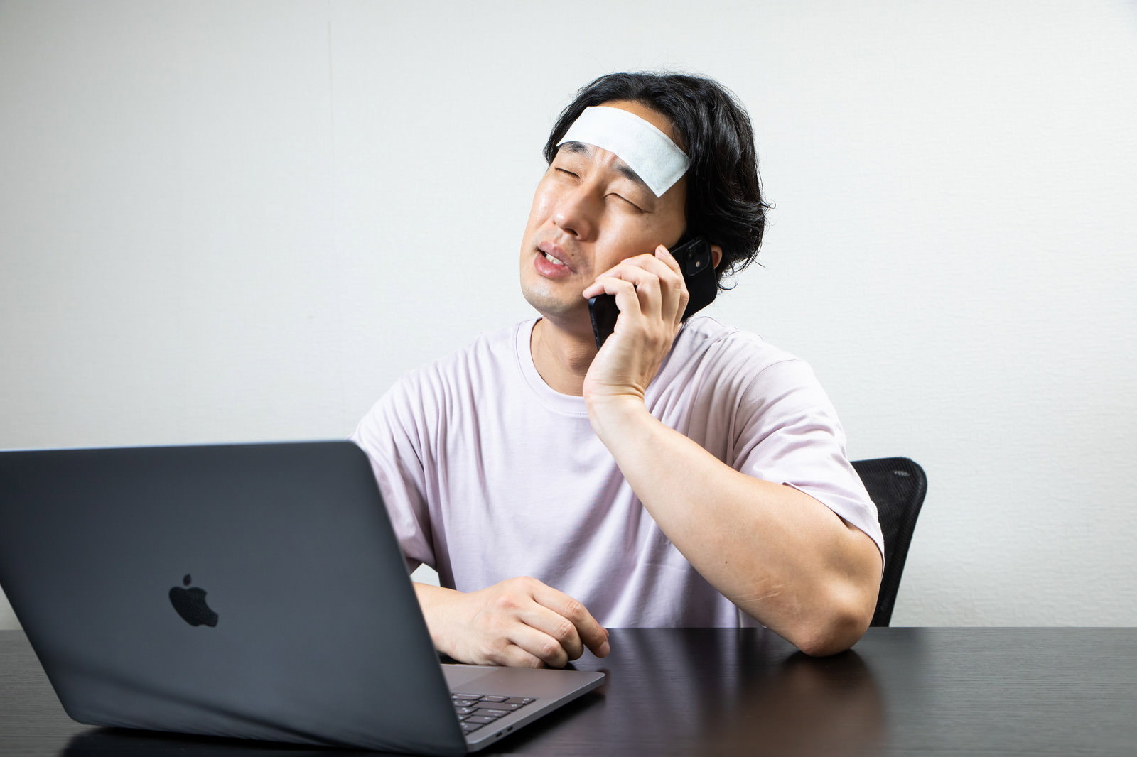 額に冷却シートを貼った男性が自宅でノートパソコンを前に電話対応している［モデル：大川竜弥］