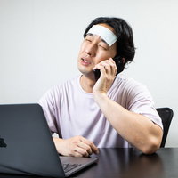 自宅でクレーム対応電話を受ける体調不良の男性、額に冷却シートの写真