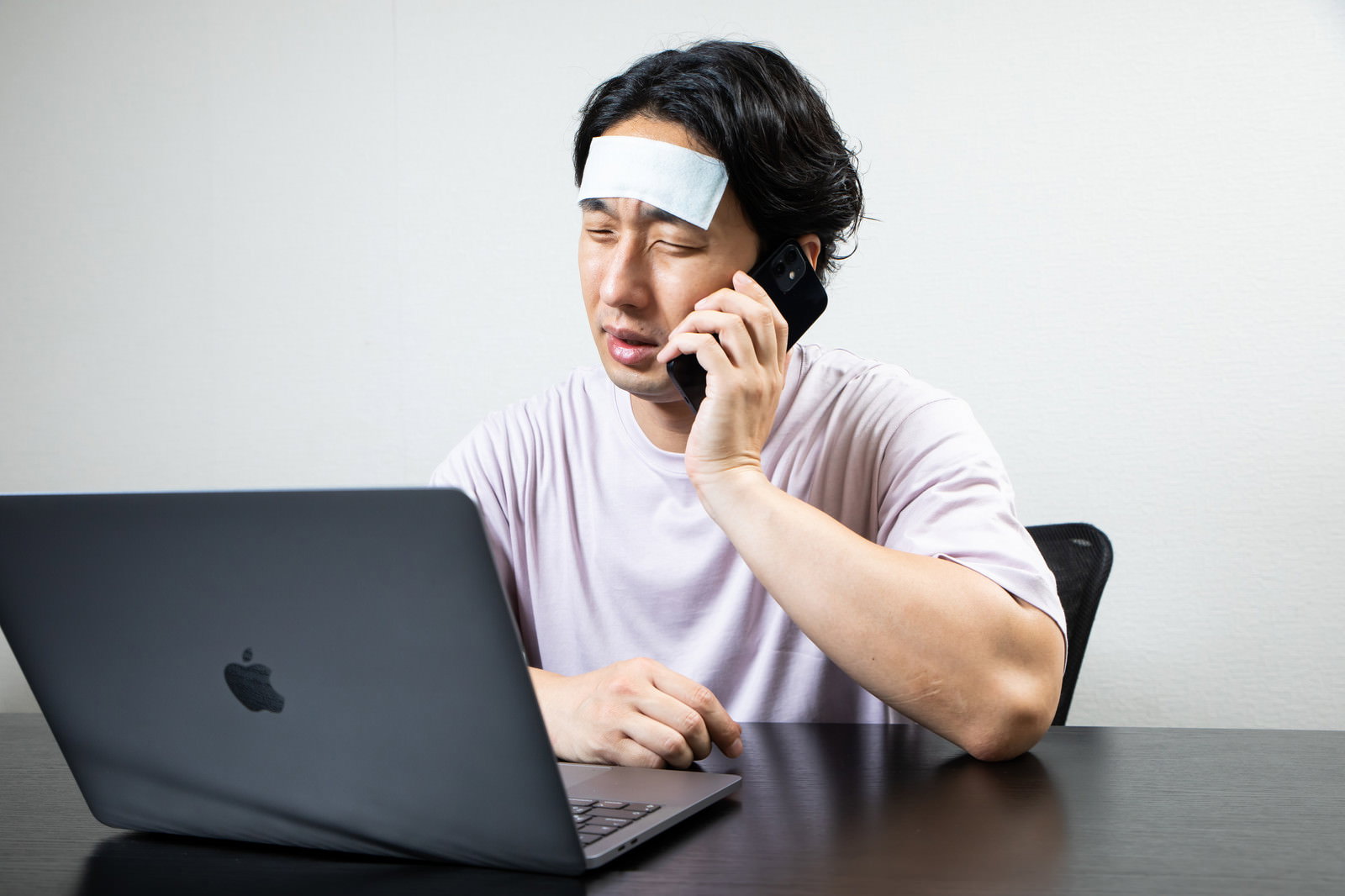 おでこに冷却シートを貼った男性がノートパソコンの前で電話をしている［モデル：大川竜弥］